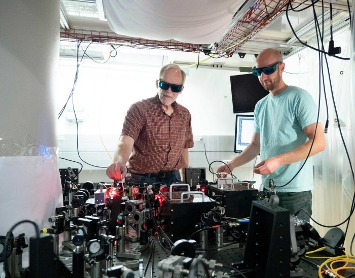 Физики NIST Джон Боллинджер (слева) и Мэтт Аффолтер настраивают лазерную и оптическую матрицу, используемую для улавливания и исследования ионов бериллия в большой магнитной камере (белая колонна слева). Ионный кристалл может помочь обнаружить загадочную темную материю. Предоставлено: Р. Якобсон / NIST.