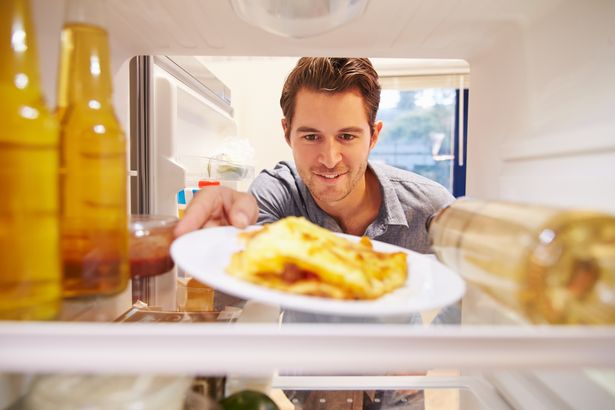 Человек смотрит внутрь холодильника, полного нездоровой пищи. Улыбается